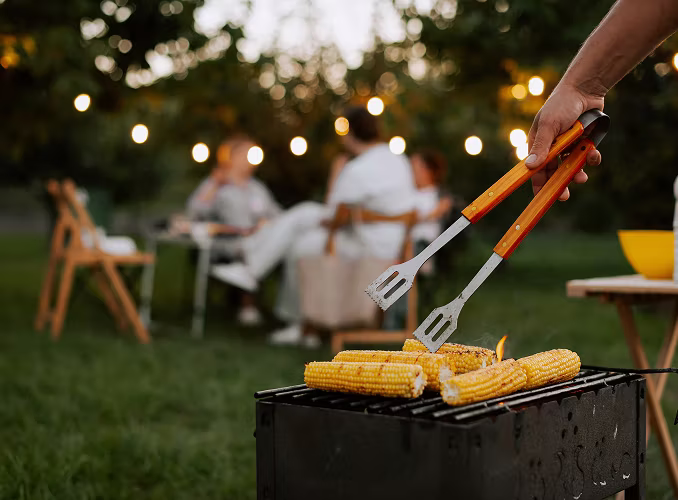 A hand turning corn cobs on a small barbecue grill in a backyard, with people seated at a table in the background under string lights.