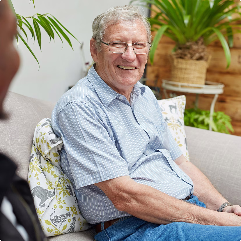 Older man sitting on the couch in a living room, speaking to another person.