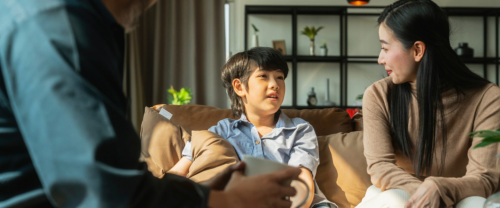 A child and a woman sitting together on a couch, talking with an older man who is seated across from them in a living room.