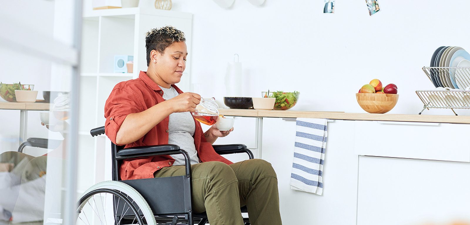A person seated in a wheelchair pouring tea from a teapot in a bright, modern kitchen with an accessible-height bench, a fruit bowl, and plants on the wall.